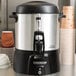 A Proctor Silex coffee urn on a counter with cups underneath the faucet.