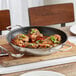 A Vigor stainless steel fry pan with meat and vegetables cooking in it on a table.