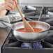 A person stirring food in a Vigor stainless steel saucier pan on a stove with a wooden spoon.