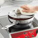 A hand holding a towel over a Vigor stainless steel saucier pan on a stove top.