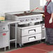 A woman in a red apron using a Cooking Performance Group Gas Countertop Charbroiler to cook hamburgers on a grill over a refrigerated chef base.