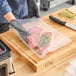 A clear vacuum packaging pouch containing raw meat and herbs on a wooden cutting board.