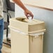 A person putting a plastic bottle into a beige Lavex slim rectangular trash can with a Drop Shot lid.