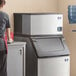 A woman standing next to a Manitowoc air cooled ice machine with a red container.