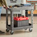 A man holding a black utility cart with gray shelves and tools on it.