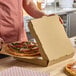 A plain kraft corrugated pizza box being used to package a pizza in a kitchen setting.