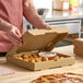 A plain kraft corrugated pizza box containing garlic knots and a dipping sauce, being opened on a kitchen counter.
