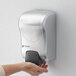 A silver San Jamar manual foam hand soap and sanitizer dispenser on a wall.