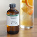 A bottle of LorAnn Oils Bergamot Super Strength Flavor next to a glass of liquid and an orange slice.