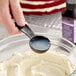 A black measuring spoon filled with liquid being held over a bowl of cream cheese mixture, with a bottle of LorAnn Oils Cream Cheese Bakery Emulsion in the background.