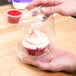 A hand holding a cupcake with white frosting and sprinkles in a clear plastic container with a hinged lid.