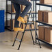 A silver and black 3-step folding step stool with a rubber hand grip being used in a warehouse setting.