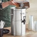 A person pouring brown liquid into a metal container with a white cylinder on top.