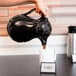 A person pouring coffee from a black Choice Thermal Swirl Coffee Carafe into a mug.