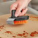 A hand using an Outset Pizza Stone cleaning brush with a scraper to clean a pizza stone.