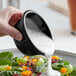 A hand pouring white sauce from a black fluted ramekin onto a bowl of salad.