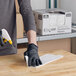 A person wearing a black glove uses a white medium-duty foodservice wiper to clean a wooden surface.