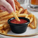 A hand holding a french fry dipping it into a black Acopa ramekin full of ketchup.