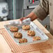 A heavy-duty quarter size aluminum sheet pan with six cookies on it, being held by a person using a towel.