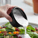 A hand pouring dressing into a bowl of salad using a black Acopa ramekin.