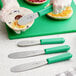A hand in a glove using a Choice stainless steel sandwich spreader with a green handle to slice a bagel.