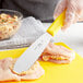 A person using a Choice Stainless Steel Sandwich Spreader with a yellow handle to spread butter on a chicken breast.