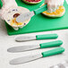 A hand in a glove using a Choice stainless steel sandwich spreader with a green handle to slice a bagel.