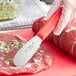 A person using a Choice stainless steel sandwich spreader with a red handle to cut meat.