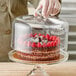 A hand holding a glass cover over a cake on a glass cake stand.