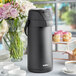 An Acopa matte black stainless steel airpot on a table with white cups of coffee.