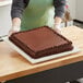 A person in gloves holding a square chocolate cake on a white wood cake board with feet.