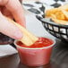 A hand holding a french fry and dipping it into a small clear plastic container of ketchup.