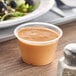 A plastic cup of sauce on a table with a bowl of food.