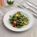 A WNA Comet white plastic plate with silver accent bands holding a salad with arugula, tomatoes, and feta cheese on a table with a glass of wine.