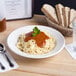 A Tuxton china pasta bowl filled with spaghetti and sauce with parsley on a table in front of a basket of bread.