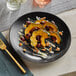 A black Acopa stoneware coupe plate with food on a table.