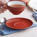 A hand holding a red and white cup of tea over a Sedona orange Acopa Keystone stoneware saucer.