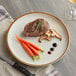A Acopa Keystone stoneware coupe plate with a steak and mushrooms on it with a knife and a glass of wine on a table.