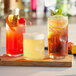 A wooden board with three Acopa beverage glasses filled with different drinks, including ice tea with a lemon slice.