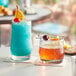 A glass of liquid next to a glass of blue liquid with a cherry on top of it.