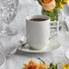 A stackable Acopa Liana bright white porcelain cup on a saucer with a brown liquid and a vase of flowers.