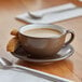 A close-up of an Acopa Embers hickory brown matte stoneware cup of coffee on a saucer with a cookie.