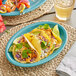 An Acopa Capri Caribbean turquoise oval stoneware coupe platter with tacos, limes, and cheese on a table.