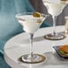 Two Acopa martini glasses with olives on top of them on a table in a cocktail bar.