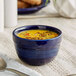 A blue Acopa Capri stoneware bouillon bowl filled with yellow soup and a spoon on a white cloth.