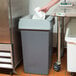 A person's hand putting a white cup in a Continental gray square trash can.