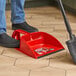 A person using a Libman red dust pan with a broom to sweep a wood floor.