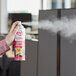 A hand holding a red and white Noble Chemical Novo aerosol can of air freshener.