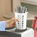 A white perforated plastic flatware holder cylinder containing metal utensils.
