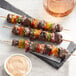 EcoChoice bamboo skewers of grilled steak and vegetables on a table next to a cup of sauce.